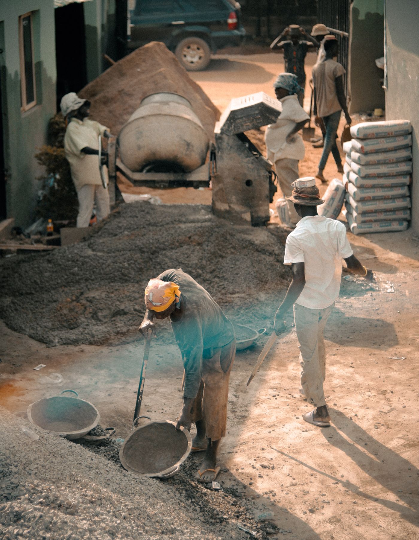 African builders on a sunny construction site loading ballast into metal basins beside a drum concrete mixer, a sand heap and a stack of cement bags