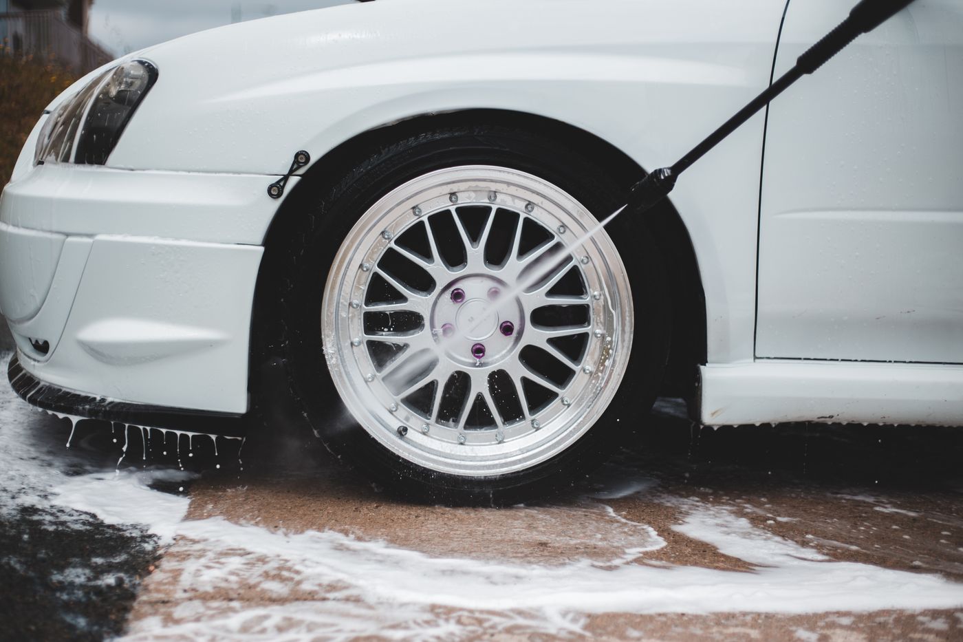 High-pressure washer lance rinsing soap suds off a car wheel at a wash bay