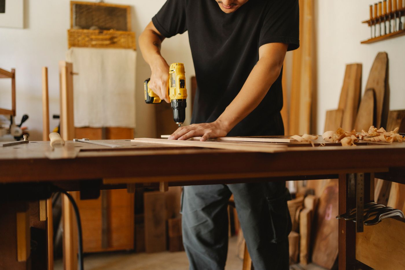 Carpenter driving a screw into a wooden plank with a yellow cordless drill on a workshop bench