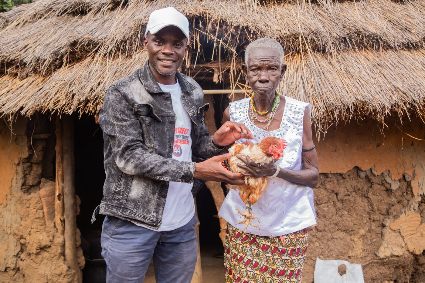 Two African poultry keepers, a younger man and an elderly woman, holding a live brown hen outside a thatched-roof rural home