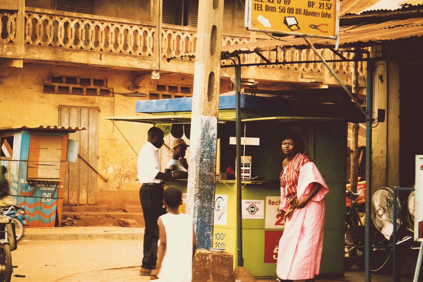 Customers at a street kiosk outside an electrical and hardware shop with a yellow sign in Abomey, Benin