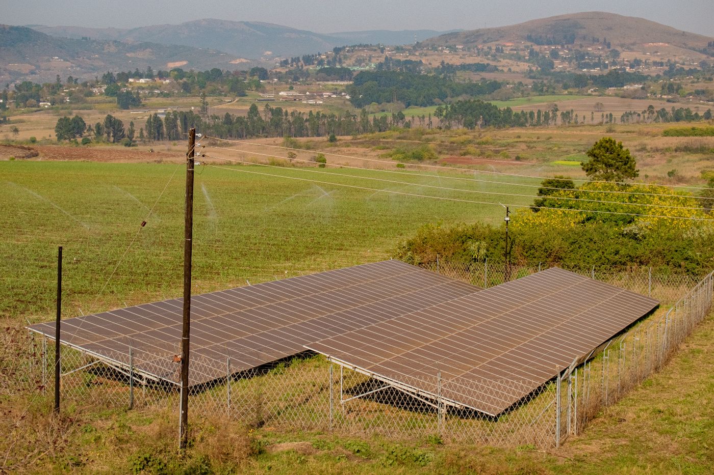 A large fenced ground-mounted solar array beside a green irrigated field with sprinklers running, power poles and hills with scattered homesteads behind