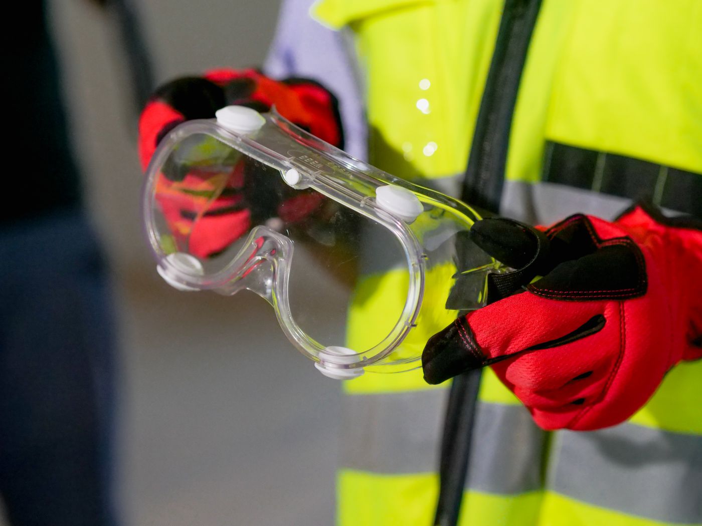 Worker in a high-visibility jacket and red work gloves holding a pair of clear vented safety goggles