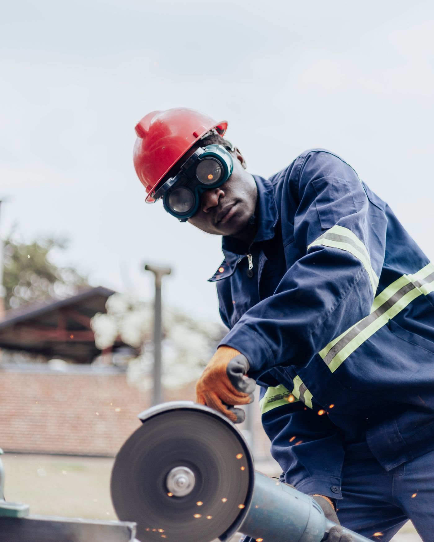An African fundi in navy overalls with reflective stripes, a red hard hat and welding goggles cutting steel outdoors with an angle grinder as small sparks fly