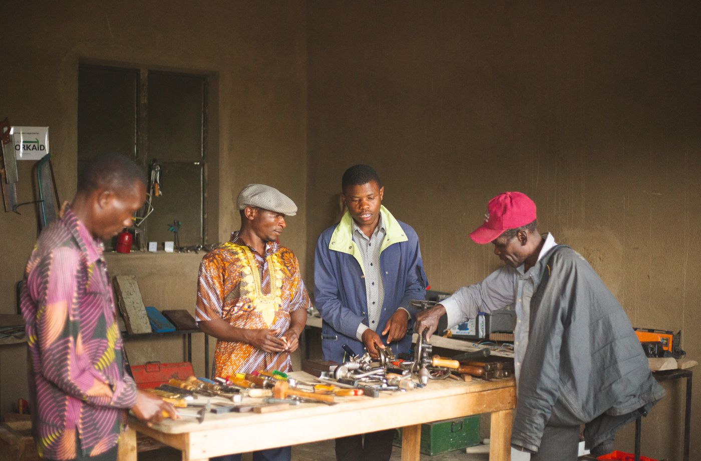 Four African men around a wooden workbench in a plastered workshop, sorting and inspecting hand drills, chisels, hammers and other hand tools laid out on the bench