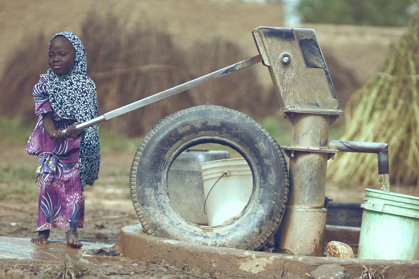 Young girl in a headscarf working the long handle of a village hand pump as water pours into a bucket