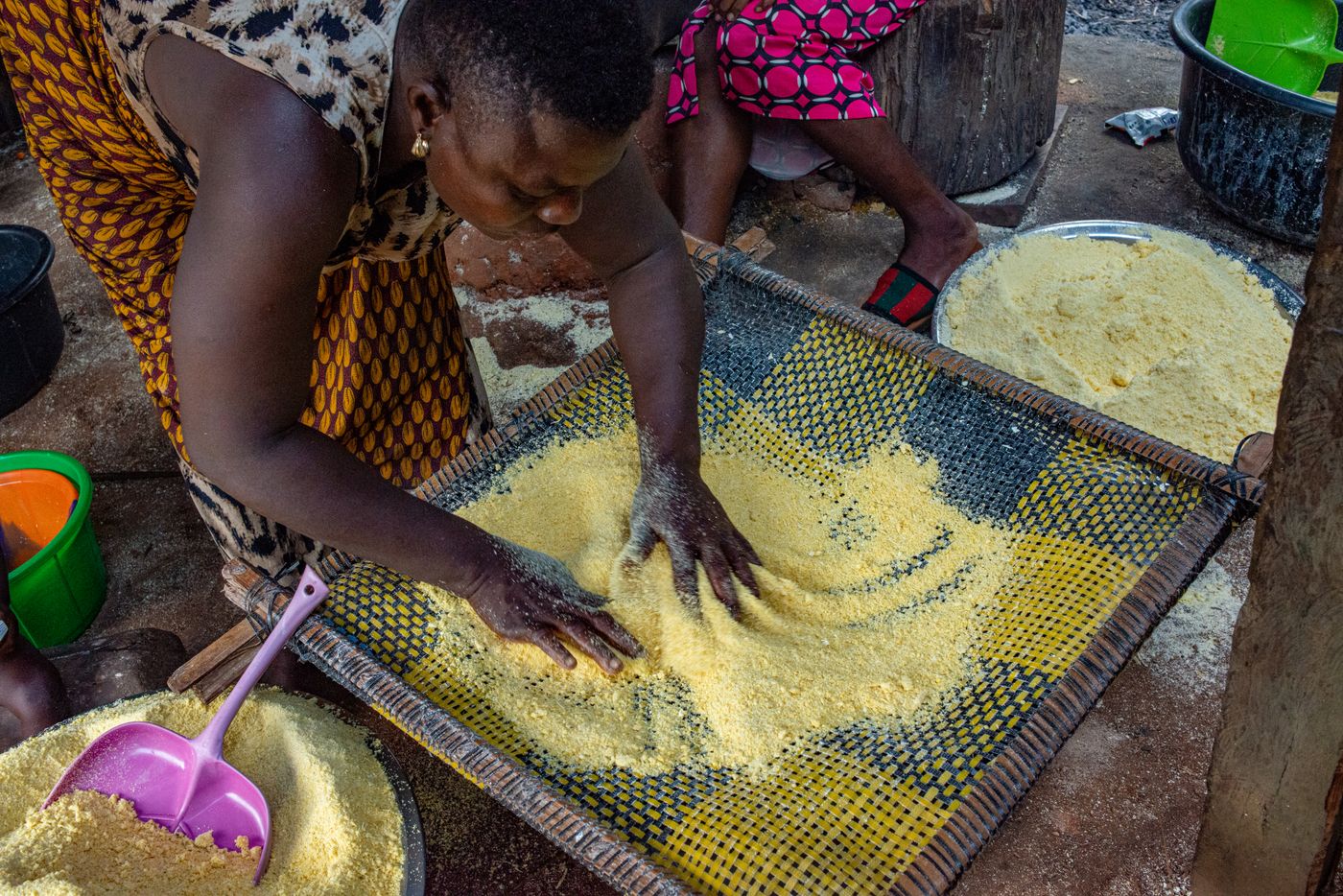 A woman shaking yellow maize meal across a woven mesh sieve in bright daylight, with bowls of sifted flour beside her