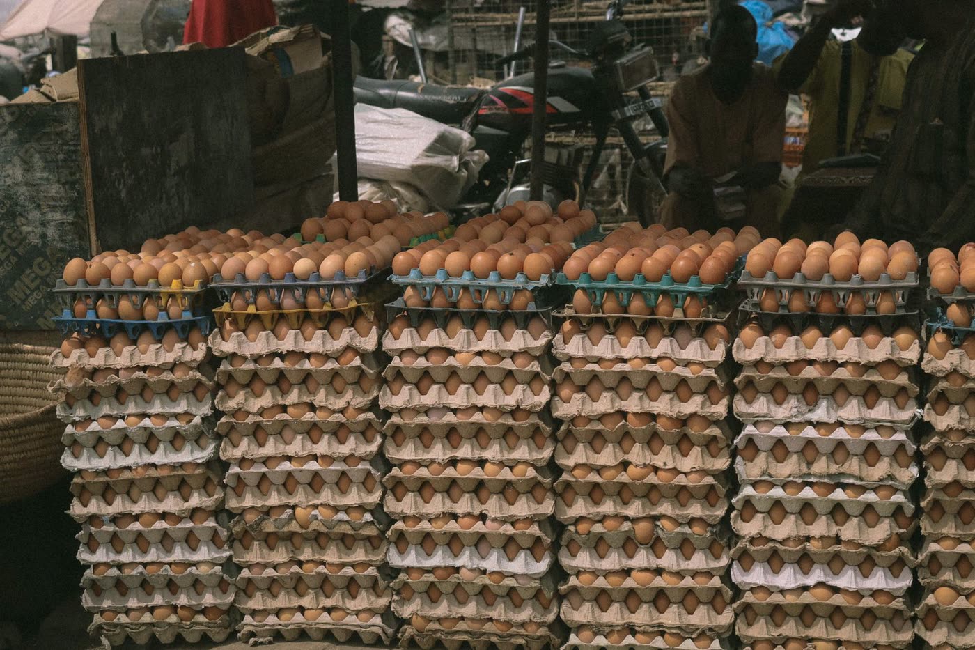Tall stacks of brown eggs in cardboard and plastic trays at an outdoor market in Kaduna, Nigeria