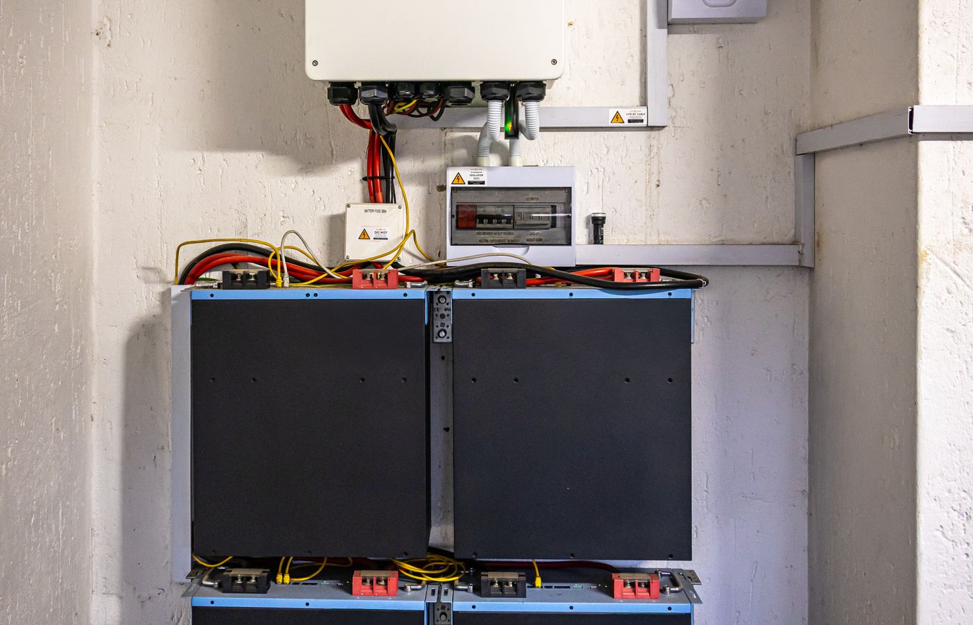 Wall-mounted hybrid inverter wired through an isolator box to a bank of lithium batteries in a bright utility room