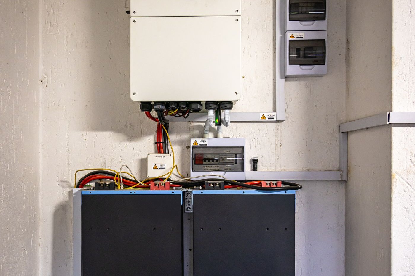 A wall-mounted hybrid inverter wired through a battery fuse and DC isolator to a bank of lithium batteries, with AC breaker boxes alongside