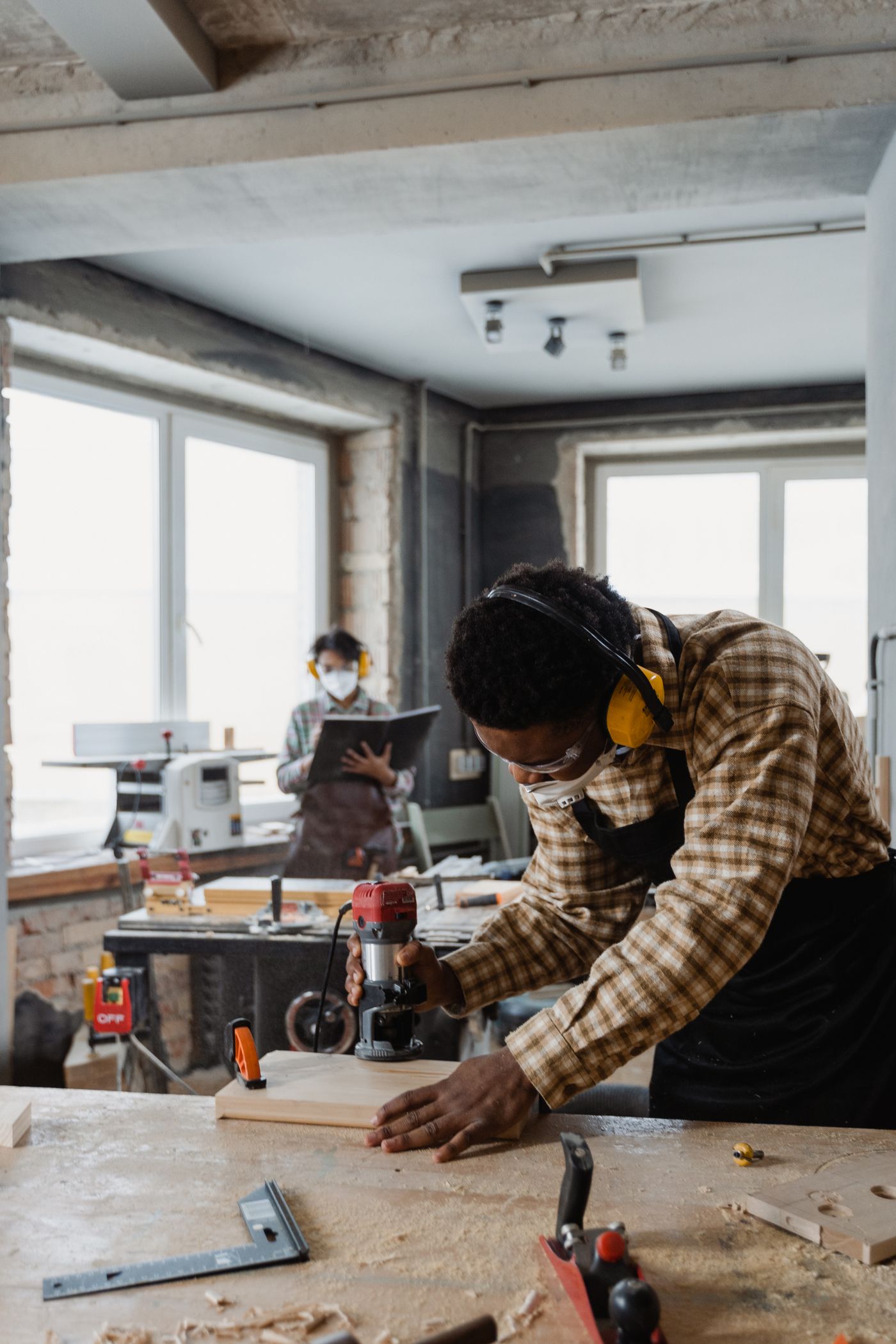 Carpenter wearing ear defenders, safety glasses and a dust mask guiding a power tool along a pine board in a joinery workshop