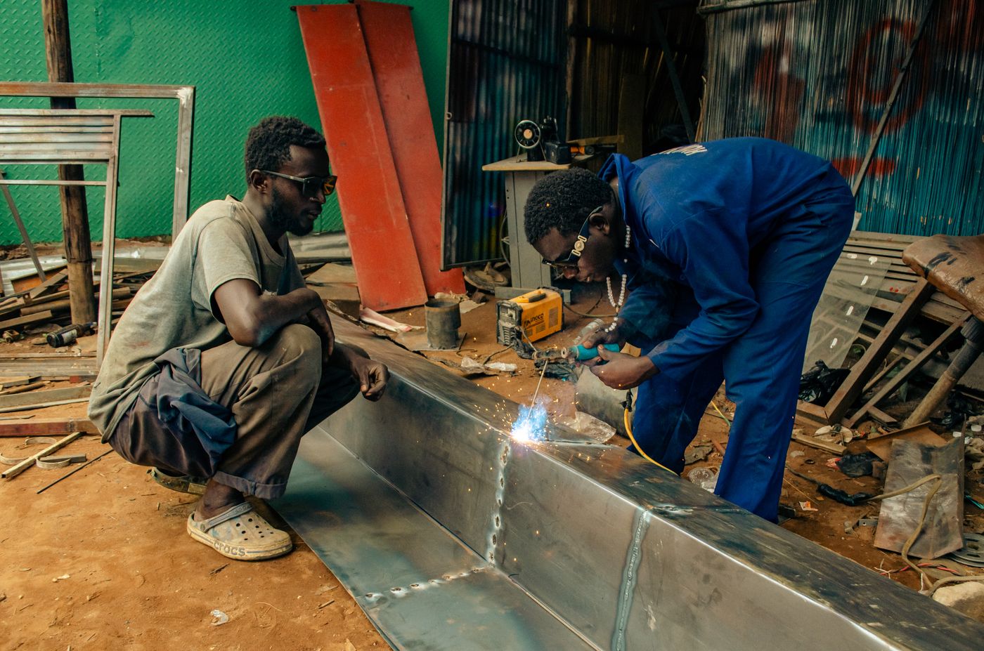 A welder in blue overalls and dark glasses arc welding a long sheet metal trough on the ground of an open workshop while a colleague crouches beside him watching, with a small portable welding machine behind