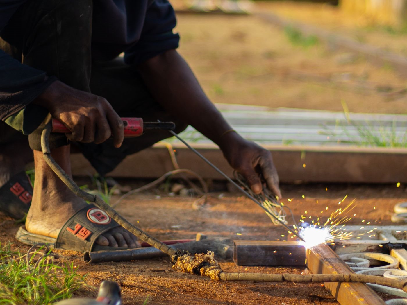 Welder crouching in an outdoor yard, striking an arc on the corner of a steel gate frame with sparks flying