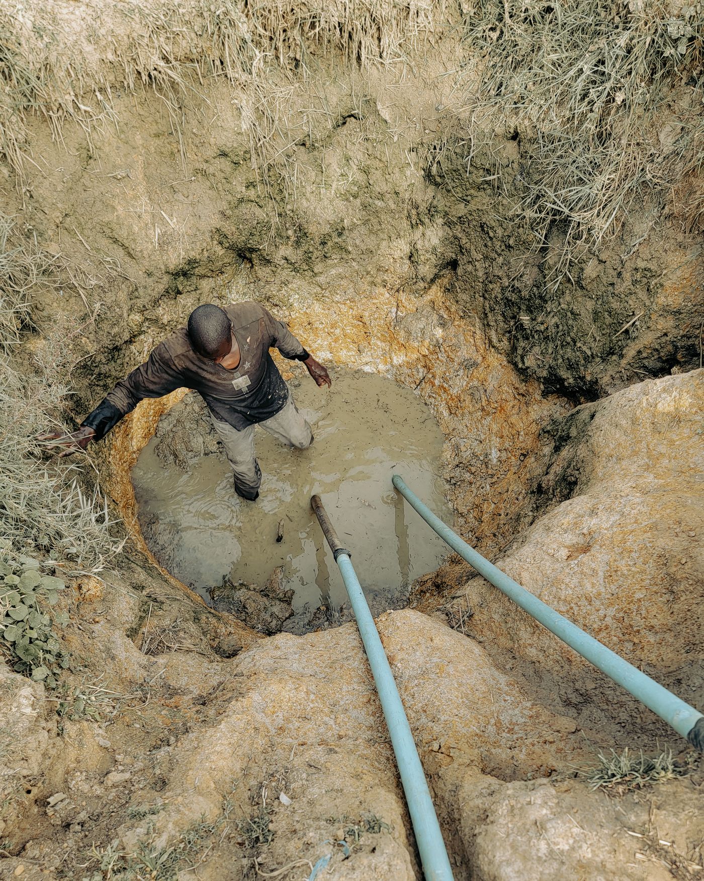 A man standing knee deep in muddy water at the bottom of a hand-dug well, with two pale blue water pipes running up the side of the pit