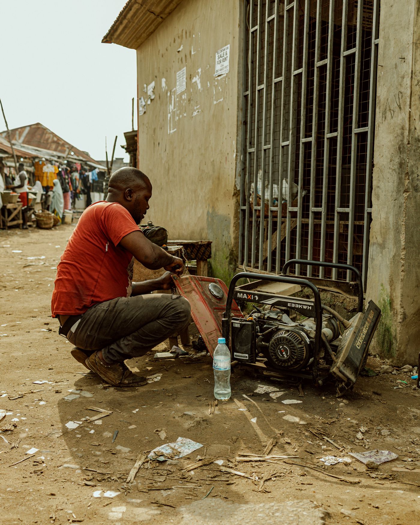A man in a red T-shirt crouching beside a portable petrol generator with its fuel tank removed, outside a shop with barred windows on a dusty market street