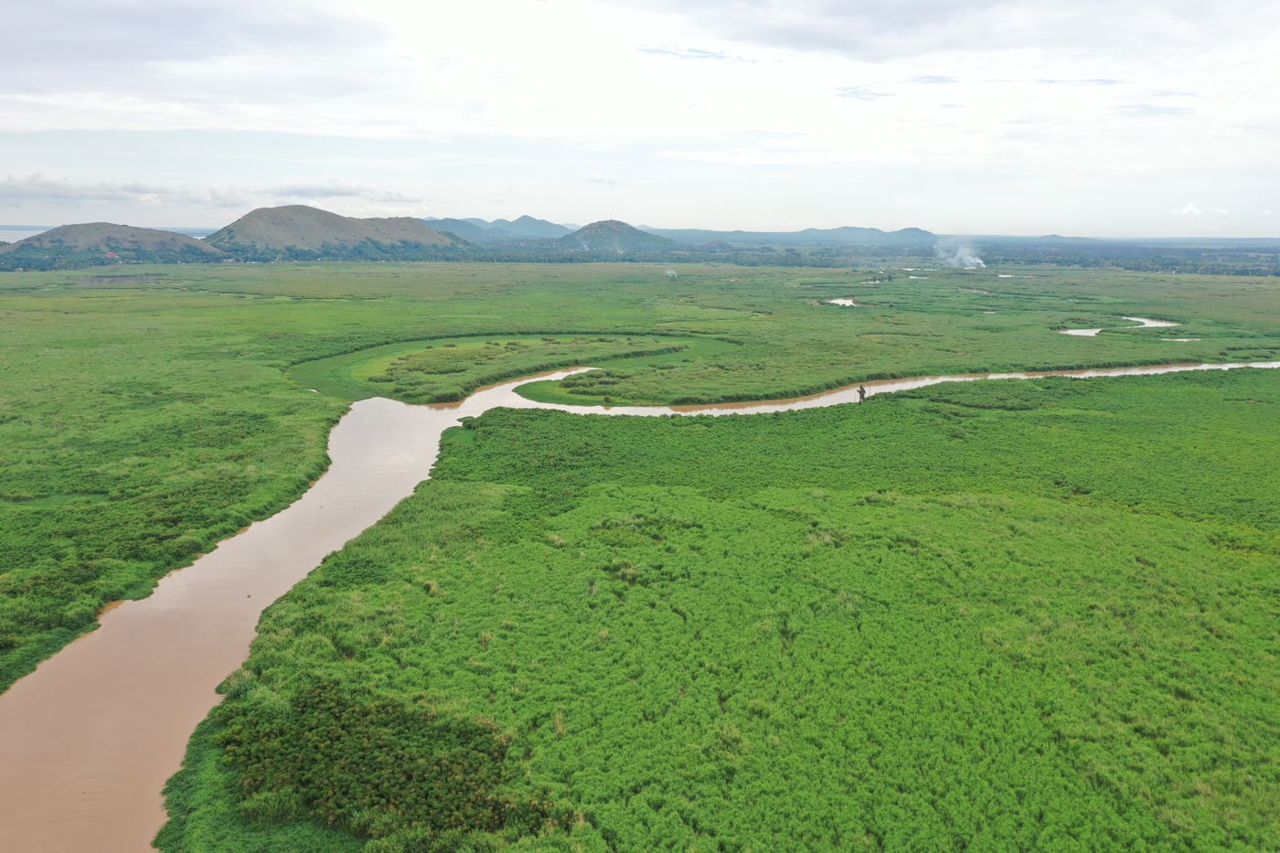 Aerial view of a brown river winding through a wide green floodplain of reeds and grass towards low hills under a cloudy sky, in Busia County, Kenya