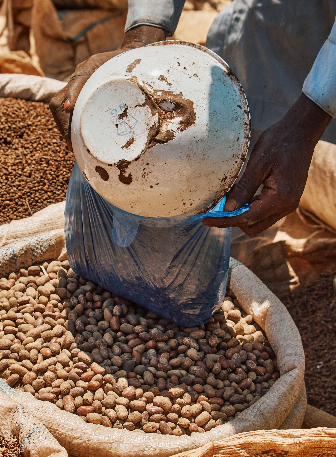 A trader's hands tipping a white enamel basin into a blue plastic bag set in an open woven sack of shelled groundnut kernels at a market