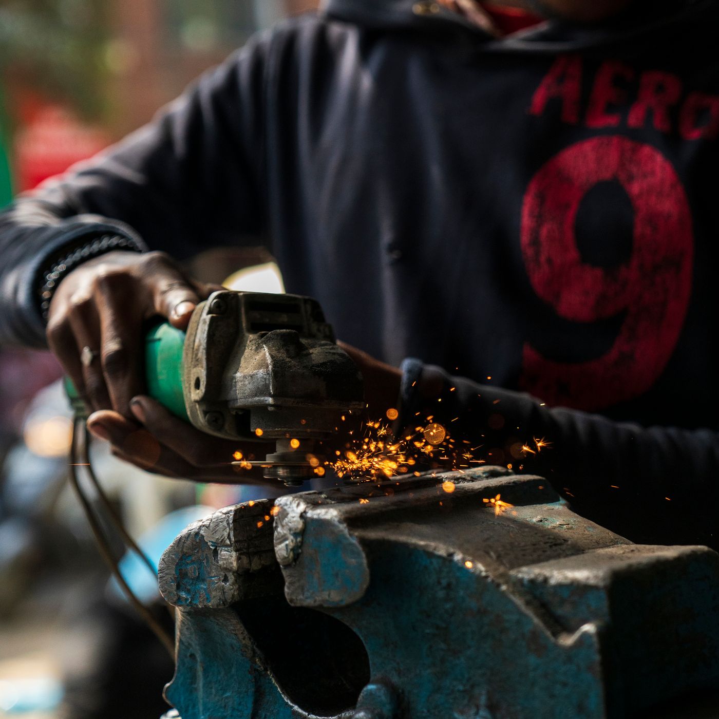 A Kenyan workshop worker using a green electric angle grinder on a blue bench vice, with bright sparks flying from the metal