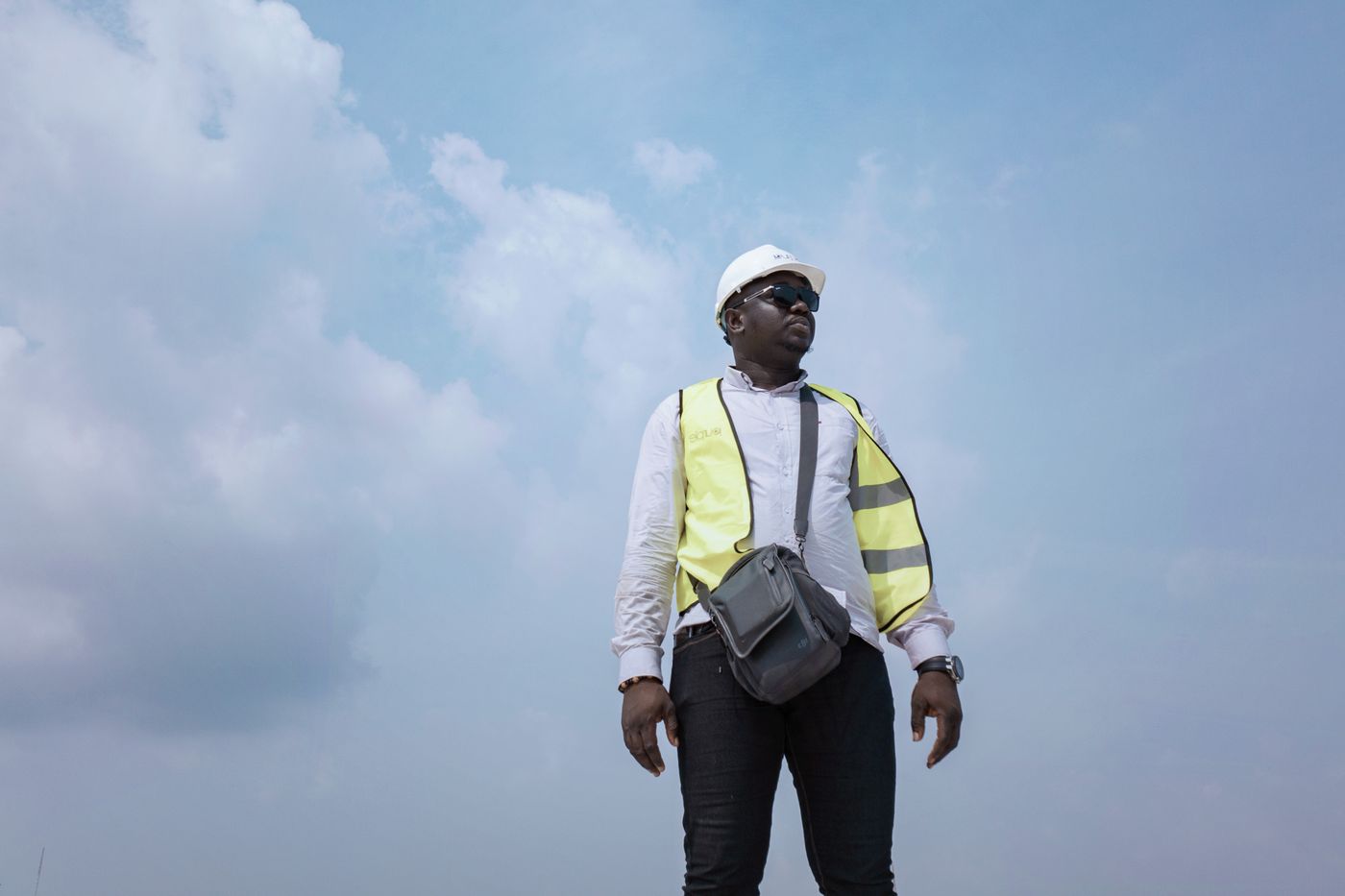 An African site supervisor in a white hard hat, sunglasses, white shirt and yellow reflective vest with a shoulder bag, standing outdoors against a pale blue sky with clouds