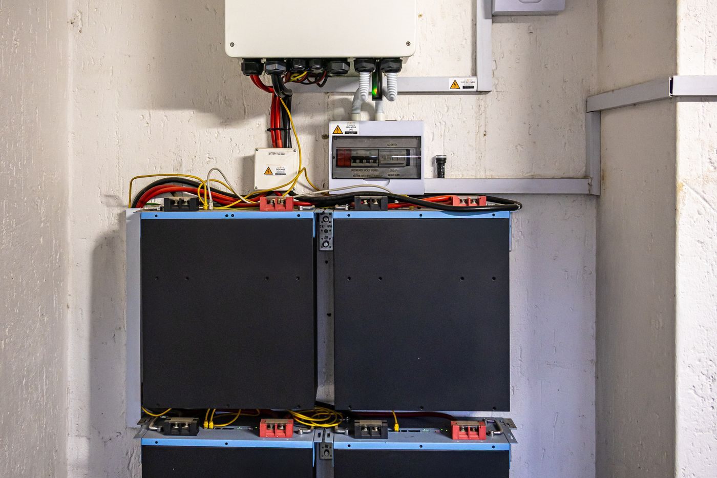Hybrid solar inverter mounted on a wall above a bank of four lithium battery modules, with a battery fuse and isolator wired between them