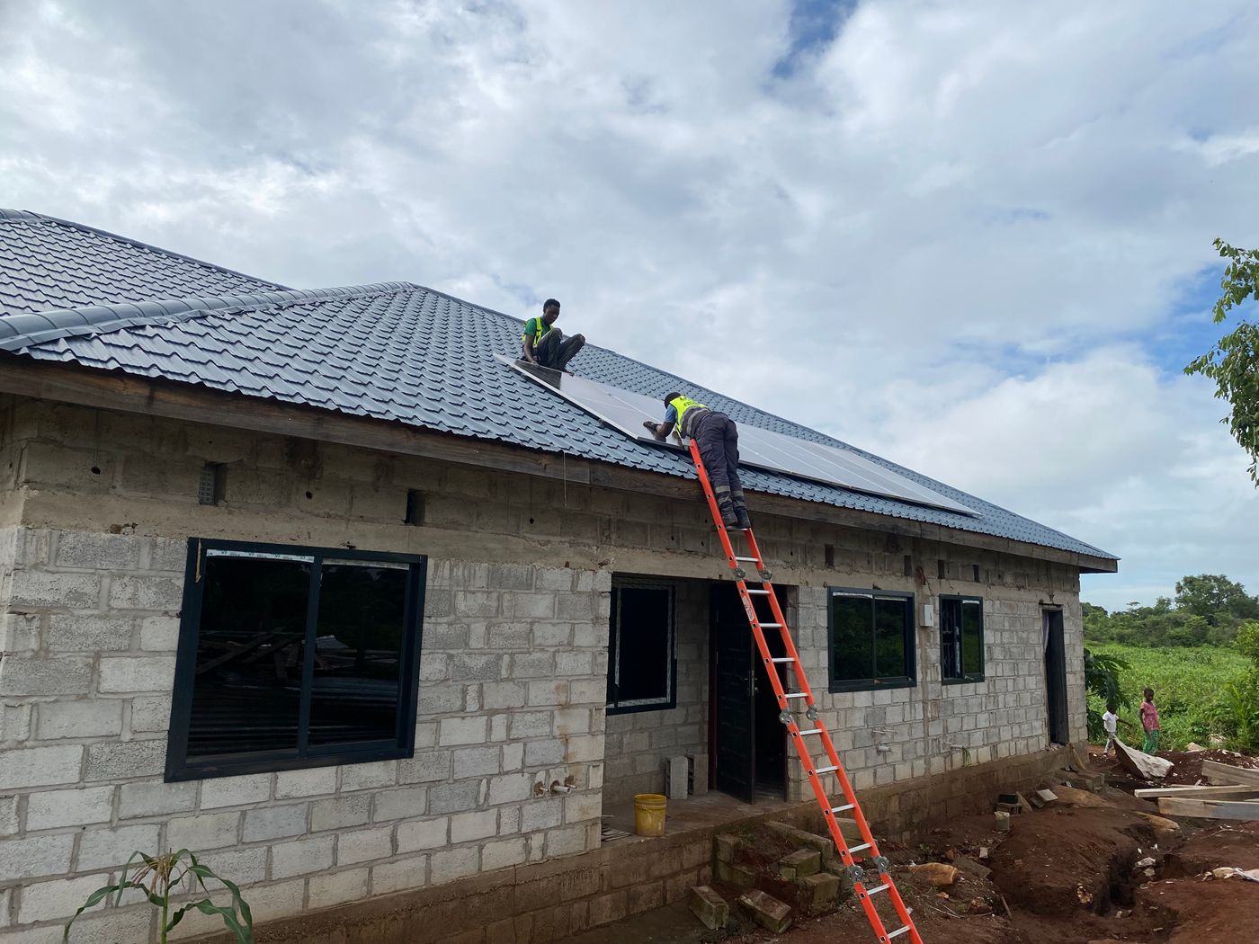 Two African installers in high-visibility vests fitting solar panels on the dark tiled metal roof of a new block farmhouse, one kneeling on the roof and one leaning from an orange ladder, with green bush and two children beside the house under a cloudy sky
