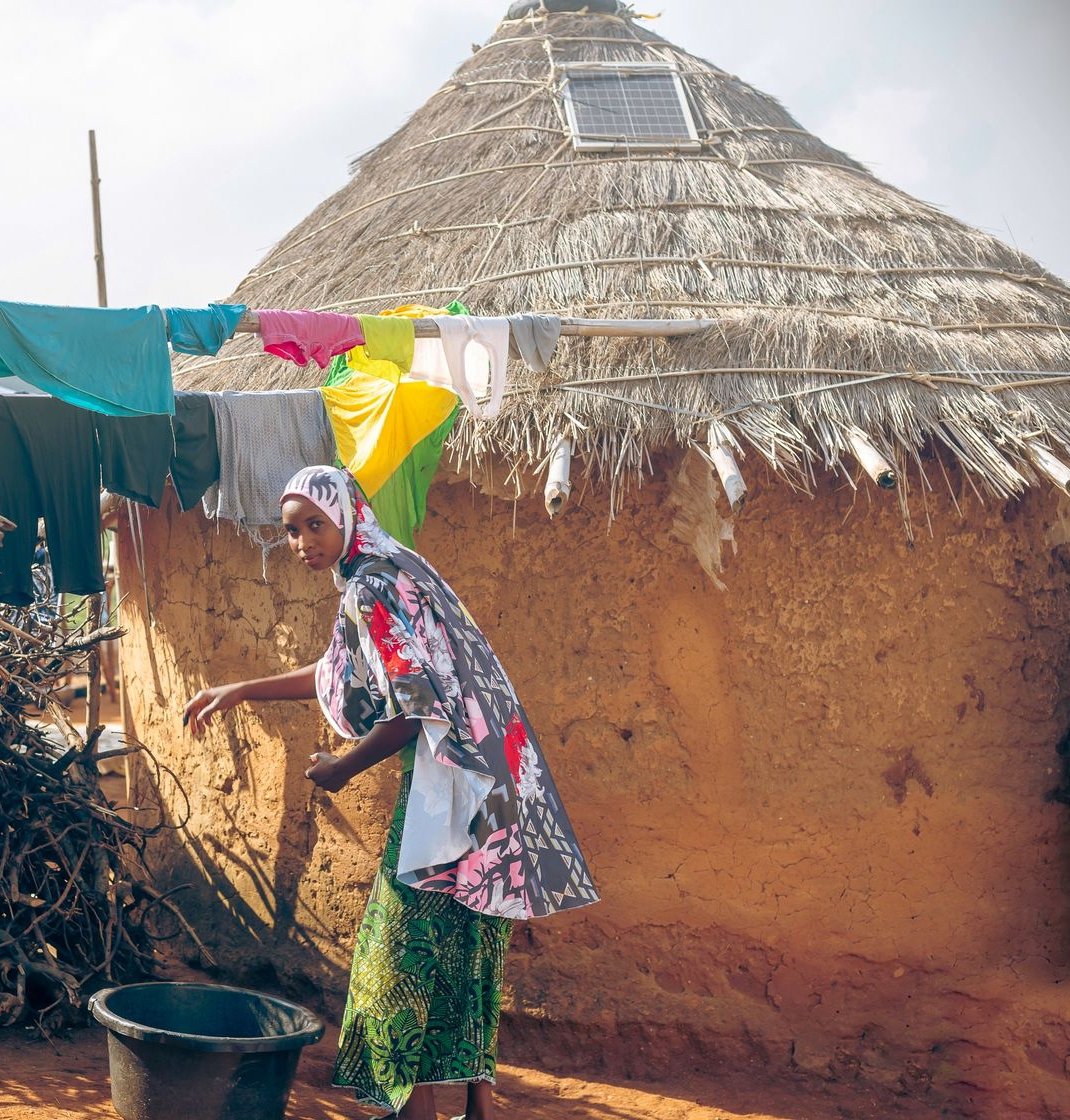 Woman in a patterned headscarf beside a mud-walled thatched hut with a small solar panel set into the roof
