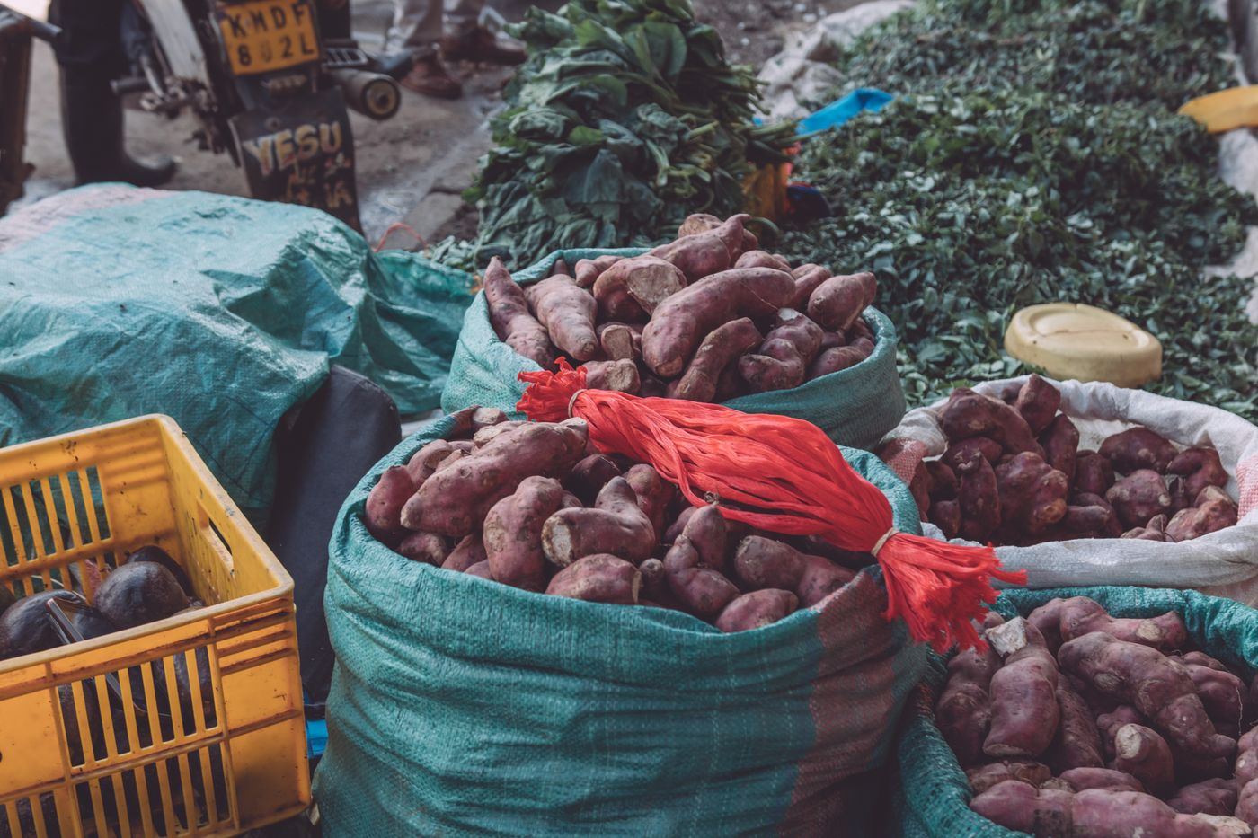 Red-skinned sweet potatoes piled in open green sacks at a street market stall in Ruaka, Kenya, beside a yellow plastic crate and bundles of leafy greens