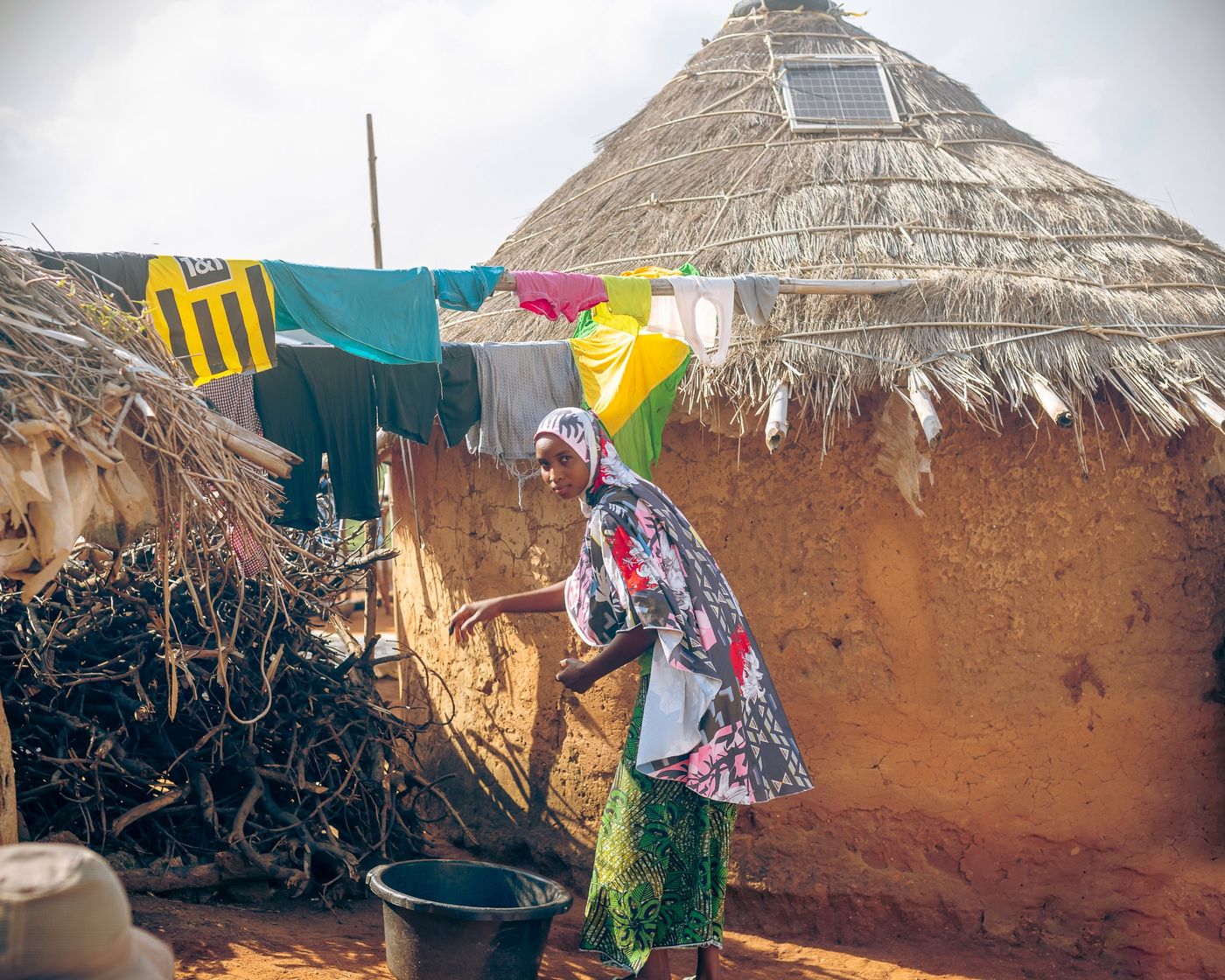 A woman in a patterned dress working outside a round thatched mud hut with a small solar panel fitted into the roof and laundry hanging on a line, in an African village