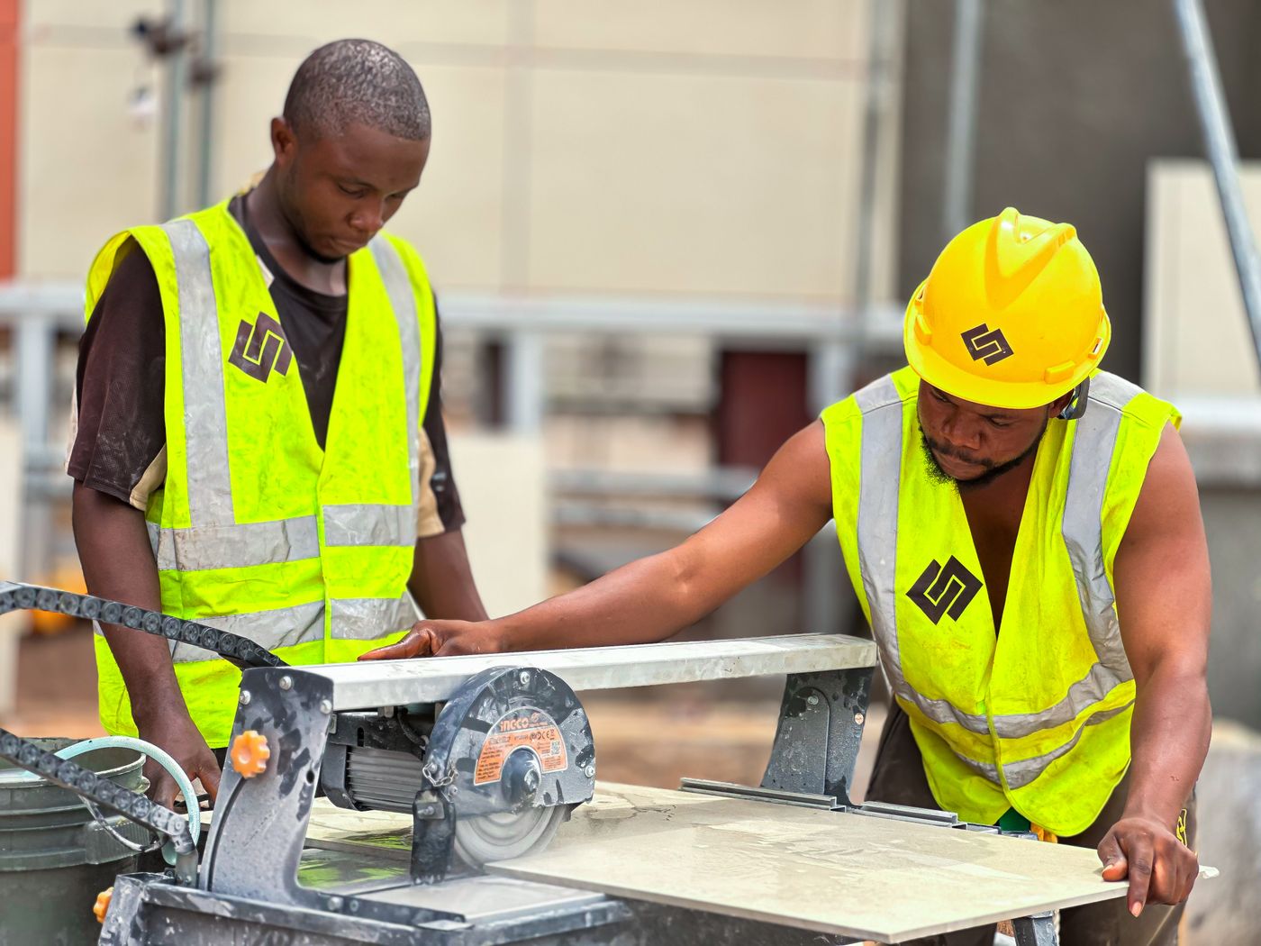 Two construction workers in high-visibility vests guiding a large porcelain floor tile through a powered tile saw on a building site