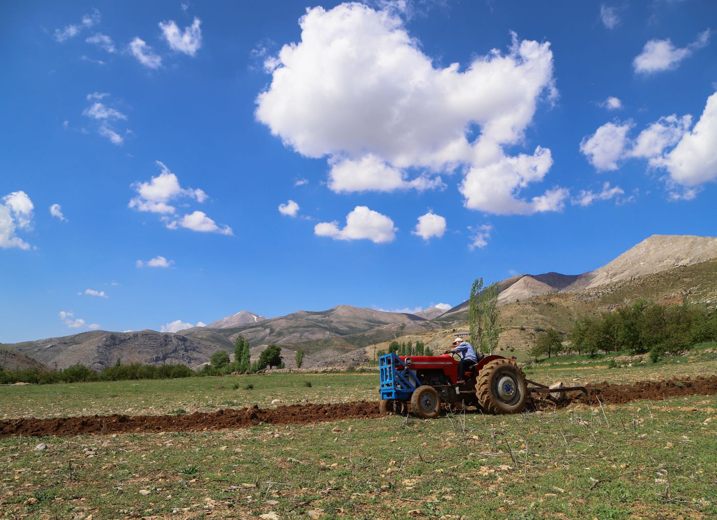 Red utility tractor pulling a mounted plough and turning a strip of fresh soil in a highland field under a blue sky