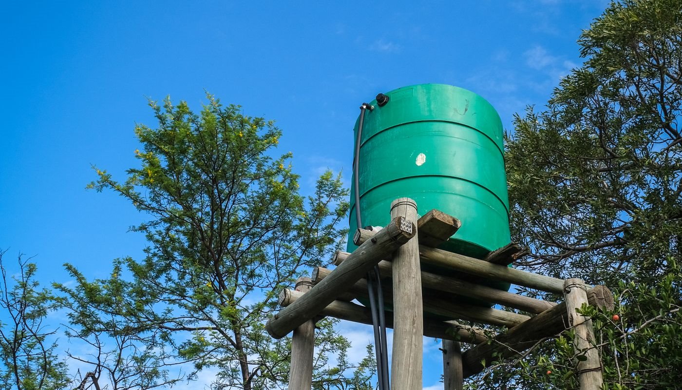 A green plastic water tank raised on a braced timber pole stand against a clear blue sky, with the supply pipe running down one leg