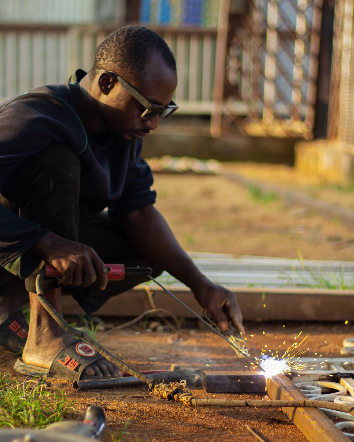 An African welder in dark glasses crouching on red soil, arc welding a decorative steel gate frame laid on the ground outdoors as sparks fly