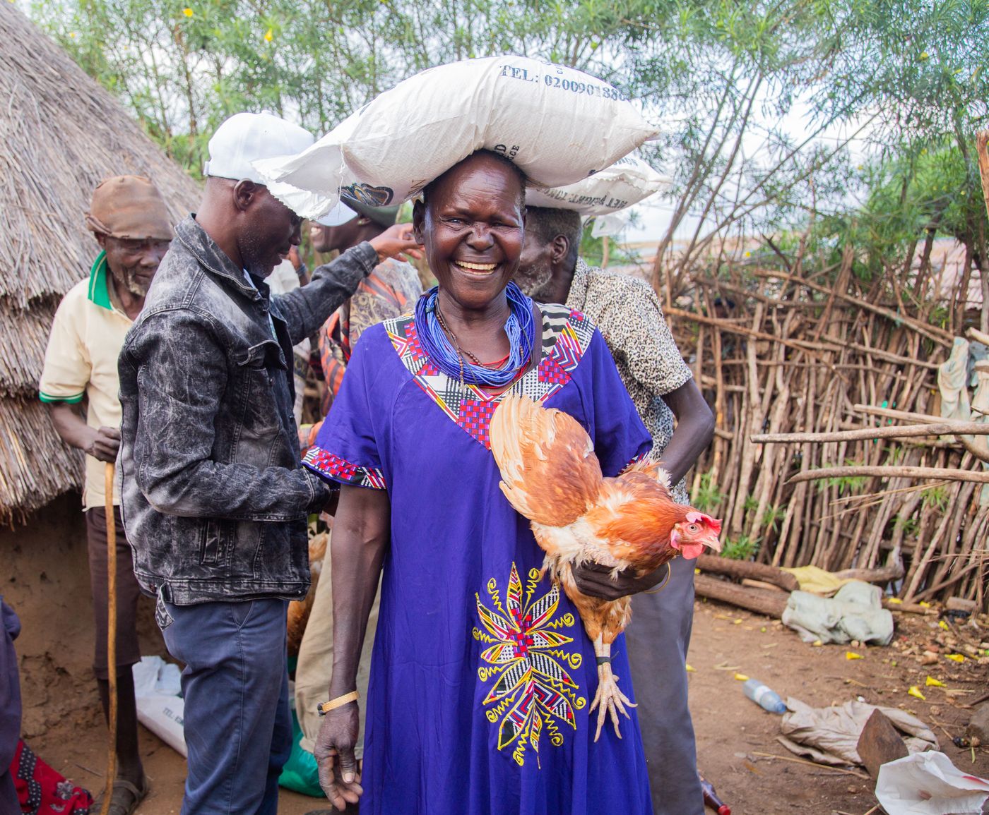 A smiling older woman in a blue embroidered dress holding a brown hen outside a mud and thatch homestead with stick fencing, as neighbours look on, in Uganda