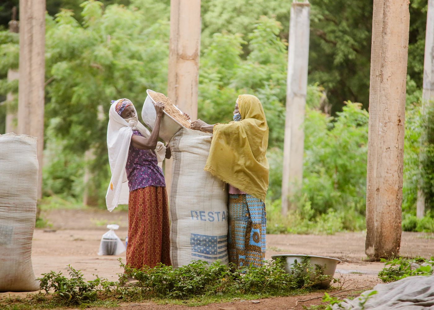 Two women in headscarves pouring grain from a metal basin into a large woven sack outdoors, with more filled sacks and concrete pillars around them, in Ghana