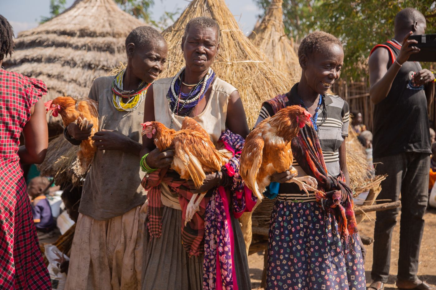 Three African women in beaded necklaces each holding a brown laying hen in front of grass-thatched granaries in a rural village