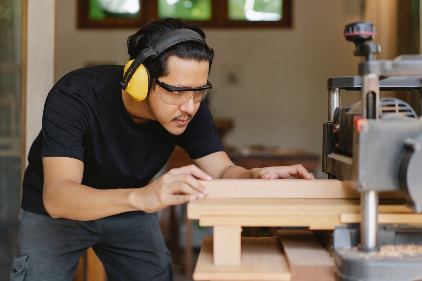 Joiner in ear defenders and safety glasses guiding a board into a bench planer, chips collecting under the outfeed